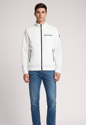 Młody mężczyzna ubrany w białą kurtkę na suwak z czarnymi detalami zamka oraz niebieskie jeansy, stojący na gładkim tle.