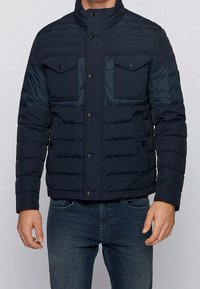 Námorníkově modrá péřová bunda s vysokým límcem, předními cvoky, dvěma hrudními kapsami a horizontálně prošívanou texturou.