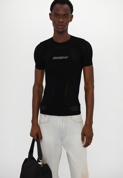Sort mesh t-shirt med kortærmer og "MISBHV" logo. Båret med lyse denimjeans og holder en sort håndtaske.
