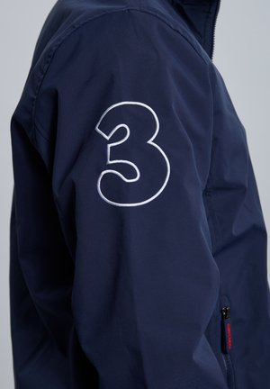 Chaqueta azul marino con un gran "3" bordado en blanco en la manga, hecha de tela suave, con cierre de cremallera en la parte delantera y bolsillos laterales.