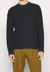 Homme portant un t-shirt noir à manches longues et un pantalon moutarde ajusté, debout devant un fond uni.