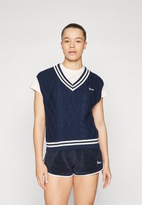 Gilet en maille marine avec un motif en losanges, bordé de rayures blanches au niveau du col en V, porté sur une chemise rose clair, associé à un short marine.