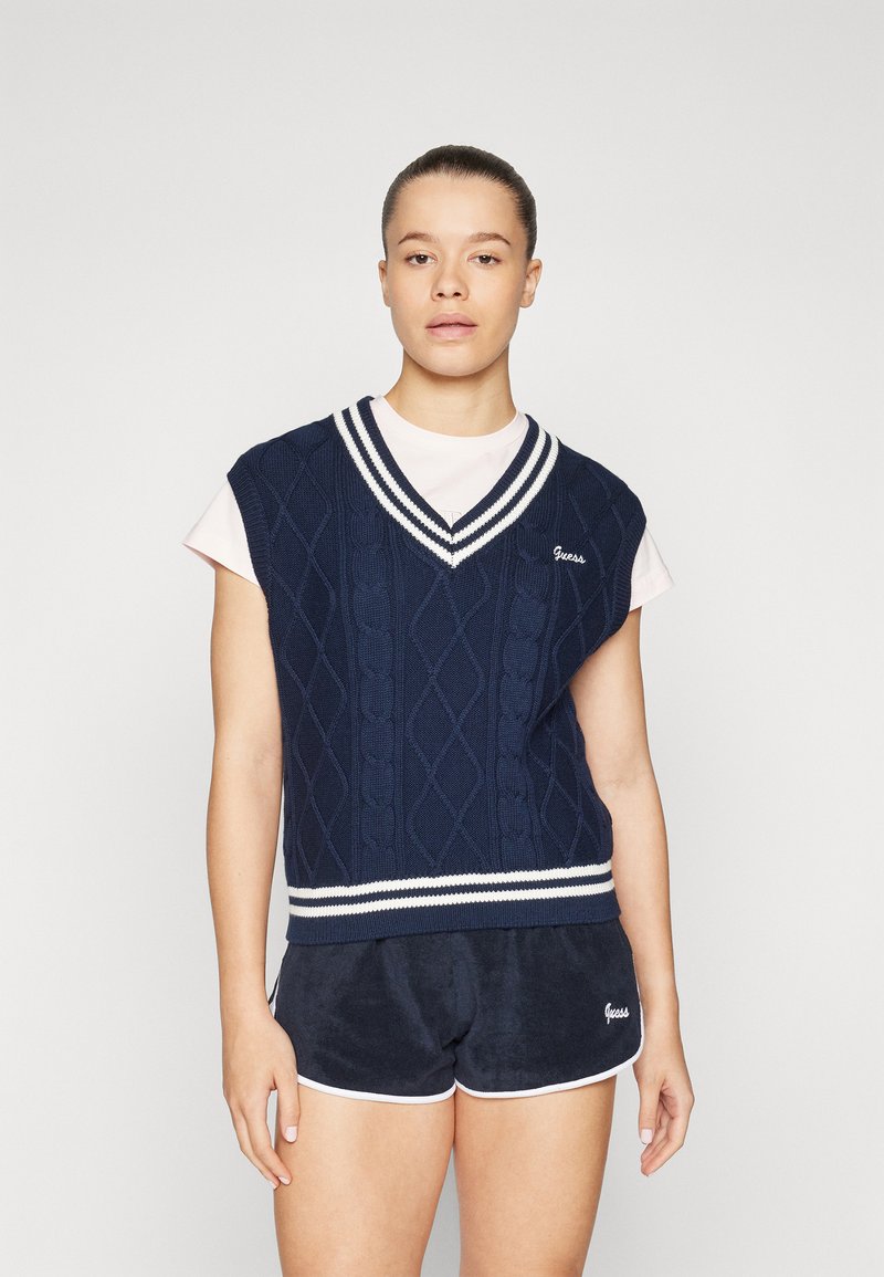 Gilet en maille marine avec un motif en losanges, bordé de rayures blanches au niveau du col en V, porté sur une chemise rose clair, associé à un short marine.