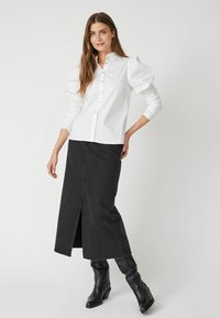 Camicia bianca con maniche a sbuffo, abbinata a una gonna di jeans nera con spacco frontale e stivali neri con tacco.