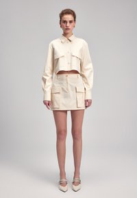 Geknipte beige blouse met knoopsluiting en klepzakken, gecombineerd met een bijpassende rok tot de knie met zijzakken en een gladde textuur.