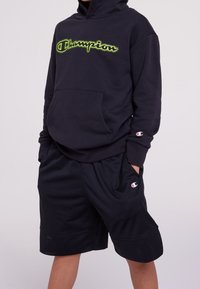 Černá mikina s kapucí s neonově zeleným logem "Champion" a přední kapsou. Kombinovaná s černými síťovanými šortkami s malým logem "Champion".