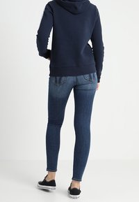 Jeans attillati blu scuro con tasche posteriori, abbinati a un hoodie blu navy e sneakers in tela nere. Tessuto morbido, design casual.