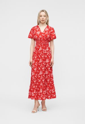 Nainen seisoo punaisessa nilkkapituisessa kukkakuvioisessa mekossa, jossa on lyhyet hihat, ja valkoiset avoimet korkokengät yksiväristä taustaa vasten.