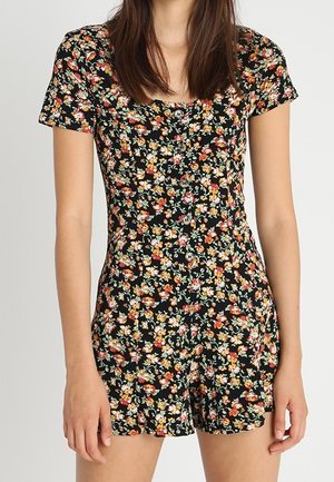 Femme portant une combinaison noire à manches courtes floral avec des boutons sur le devant et des fleurs rouges, jaunes et blanches.