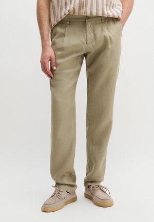 Mann trägt beige gerade geschnittene Hosen, ein in die Hose gestecktes gestreiftes Kurzarmhemd und hellbraune, geschnürte Freizeitschuhe, steht mit den Händen in den Taschen.