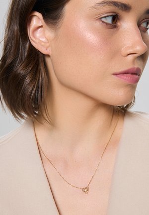 Złoty naszyjnik z sercowym wisiorkiem, ozdobionym kwiecistymi detalami, noszony na jasnym topie. Gładka tekstura i delikatny wzór łańcuszka.