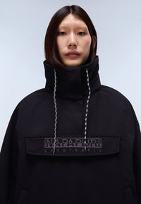 Joven con cabello lacio y oscuro que lleva una chaqueta negra de cuello alto con cordones y un gran panel con el logo "Napapijri geographic".