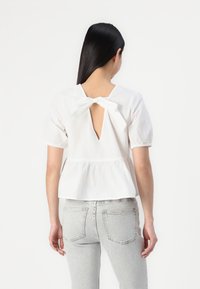 Fehér pamut felső puffos ujjal, hátul kulcslyuk részlettel és masnival. Peplum szegéllyel és sima textúrával rendelkezik. Világosszürke farmernadrággal párosítva.