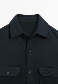 Mørkeblå skjorte med nedslået krave, to brystlommer og knaplukninger. Stoffet fremstår struktureret og slidstærkt.