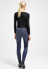 Jeans oscuros ajustados de color azul con un diseño clásico de cinco bolsillos, ligero desgaste y costuras decorativas en los bolsillos traseros. Combinados con una blusa negra de manga larga.