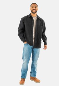 Camisa negra de botones con dos bolsillos frontales, llevada sobre una camiseta gris. Jeans azules y botas marrones completan el atuendo. Fondo blanco liso.