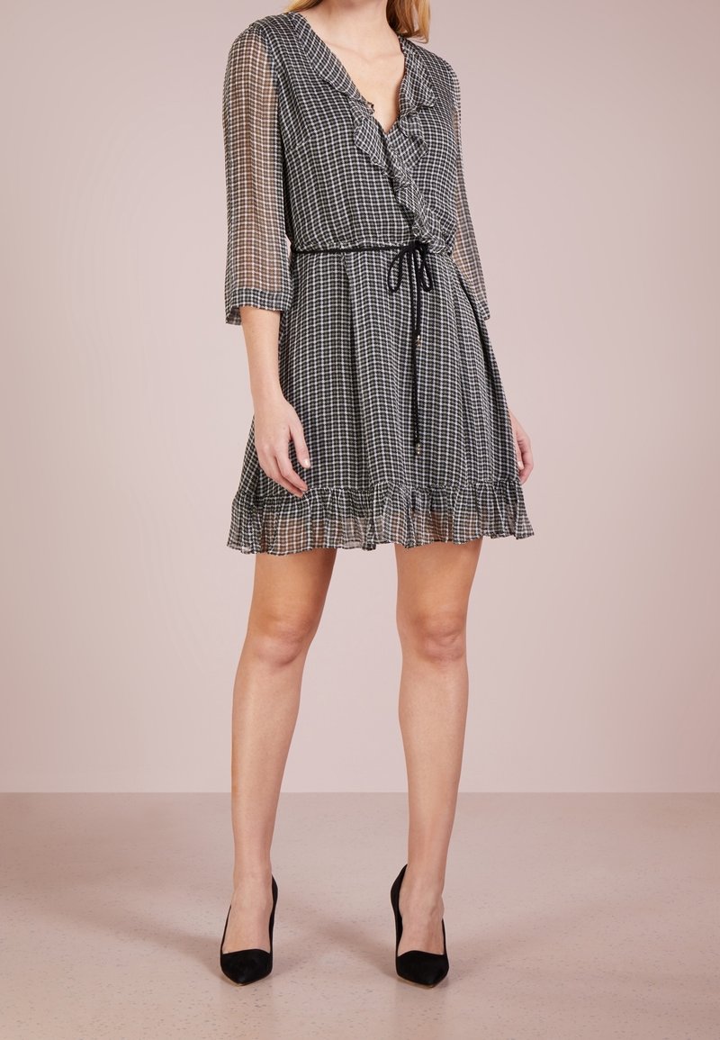 Femme portant une robe à carreaux noir et blanc avec un décolleté en V à volants, des manches transparentes, une ceinture à nouer à la taille et des chaussures à talons noirs.