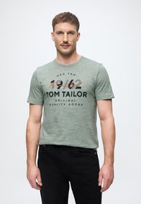 Πράσινο βαμβακερό t-shirt με χρώμα heather, κοντά μανίκια, στρογγυλή λαιμόκοψη, με σχέδιο γραφικών κειμένων και λογότυπο σε μαύρο και πορτοκαλί χρώμα στο μπροστινό μέρος.