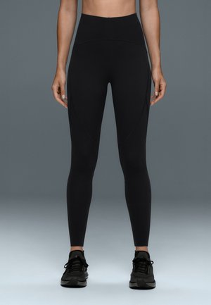 Vue de face d'une personne portant des leggings noirs taille haute et des chaussures de sport noires, debout avec les bras détendus le long du corps.