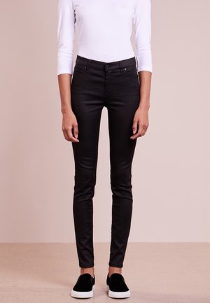 Person iført sorte skinny bukser, hvid langærmet top og sorte slip-on sneakers stående mod ensfarvet baggrund.