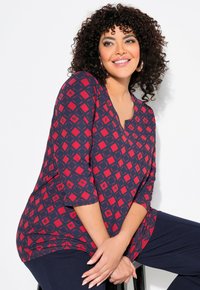 Top rosso e blu con motivo a V e maniche a tre quarti, caratterizzato da forme geometriche e un tessuto morbido. Abbinato a pantaloni scuri.