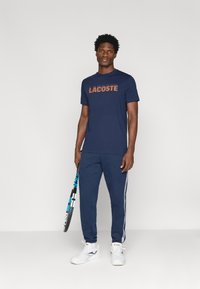 Ναυτικό βαμβακερό μπλουζάκι με την εγγραφή "LACOSTE" σε καφέ γράμματα, συνδυασμένο με ναυτικές φόρμες που διαθέτουν λευκές πλευρικές ρίγες και λευκά αθλητικά παπούτσια.