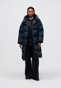 Námořnický péřový kabát s dlouhým střihem, velkými prošívanými sekcemi a prostornou kapucí. V kombinaci s tmavým trikem a kalhotami s širokými nohavicemi.