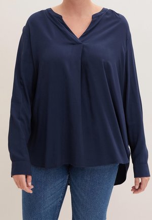 Person trägt eine locker sitzende, marineblaue Langarmbluse mit V-Ausschnitt und blaue Jeans vor einem einfarbigen Hintergrund.