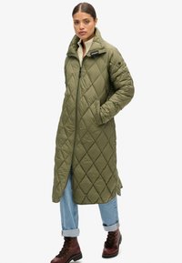 Manteau long matelassé vert avec un col haut et une fermeture éclair à l'avant ; avec des poches latérales et une coupe décontractée ; porté sur un jean bleu clair et des bottes marron.