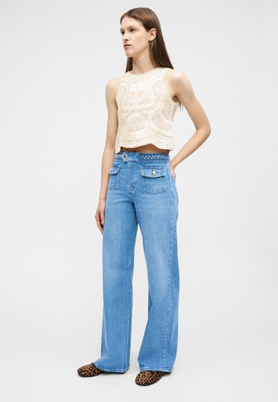 Γυναίκα που στέκεται φορώντας μπεζ αμάνικο πλεκτό τοπ, ψηλόμεσα μπλε τζιν με μπροστινές τσέπες και παπούτσια με λεοπάρ μοτίβο.