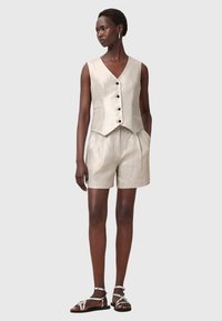 Linnenväst med v-ringning och svarta knappar, ihop med matchande linneshorts. Sandaler med silverfärgade remmar. Neutral ljusbeige färg.