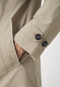 Beige-aines ulkovaatteet, joissa on sileä pinta; tummat napit, joissa on kontrastivärinen ommel, ja hihan sivulla näkyvä tasku.