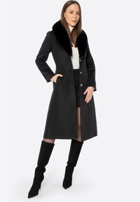 Czarny płaszcz z dużym, miękkim, czarnym futrzanym kołnierzem, o dopasowanym kroju i złotych guzikach; noszony na białej bluzce i zestawiony z czarnymi kozakami sięgającymi kolan.