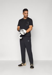 Black athletic t-shirt with a chest pocket, black pants with a logo, and black and tan shoes. The model holds white boxing gloves.