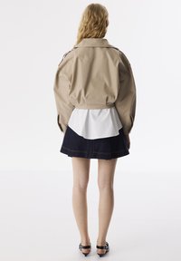 Beige Cropped-Jacke mit hohem Kragen, getragen über einem weißen Peplum-Shirt, kombiniert mit einem dunklen Jeansrock und schwarzen Absatzsandalen.