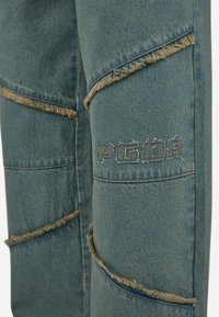 Blauwe denim broek met rafelige randen, patchworkontwerp en geborduurd "PGDR" logo. De stof heeft een gestructureerde, versleten uitstraling.