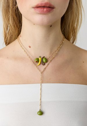Približnji posnetek ženske vratu z večplastnimi zlato verižicami z obeski v obliki zelenih oljk, rumenih čilijev in rjavih fižolov, čez belo majico.