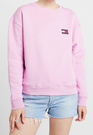 Sweatshirt rose avec un col rond, des poignets et un ourlet côtelés. Présente un petit patch logo Tommy Jeans sur la poitrine gauche. Porté avec un short en denim.
