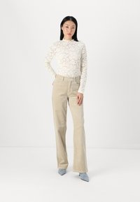 Bijela čipkasta bluzica dugih rukava s cvjetnim uzorkom, u kombinaciji s bež hamsterm pants. Model nosi svijetloplave šiljate štikle.