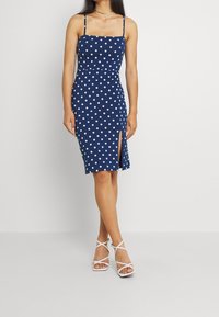 Femme portant une robe midi sans manches à pois blancs sur fond bleu marine, avec une fente frontale, associée à des sandales blanches à lanières et des colliers en or.