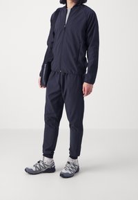 Muž v černé sportovní bundě a kalhotách s bílými sportovními botami a ponožkami, drží černou tašku přes rameno a stojí na bílém podlaze.