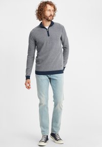 Pulover cu fermoar de culoare bleumarin și gri, cu un model texturat, având guler în picioare, asortat cu blugi albaștri deschiși și adidași negri.