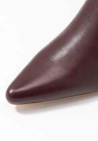 Chaussure à talon haut en cuir bordeaux avec un bout pointu, une texture lisse et une semelle beige clair. Pas de motifs ni d'éléments métalliques visibles.