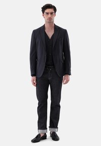 Dunkler Slim-Fit-Blazer über einem schwarzen Hemd mit Knopfverschluss, kombiniert mit dunklen Jeans. Schwarze Slipper runden den Look ab. Einfarbiger Hintergrund.
