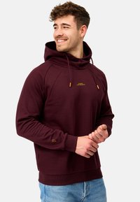 Haut à capuche bordeaux avec cordon de serrage, poignets côtelés et un petit logo doré sur la poitrine. Texture lisse, coupe décontractée et poche kangourou.
