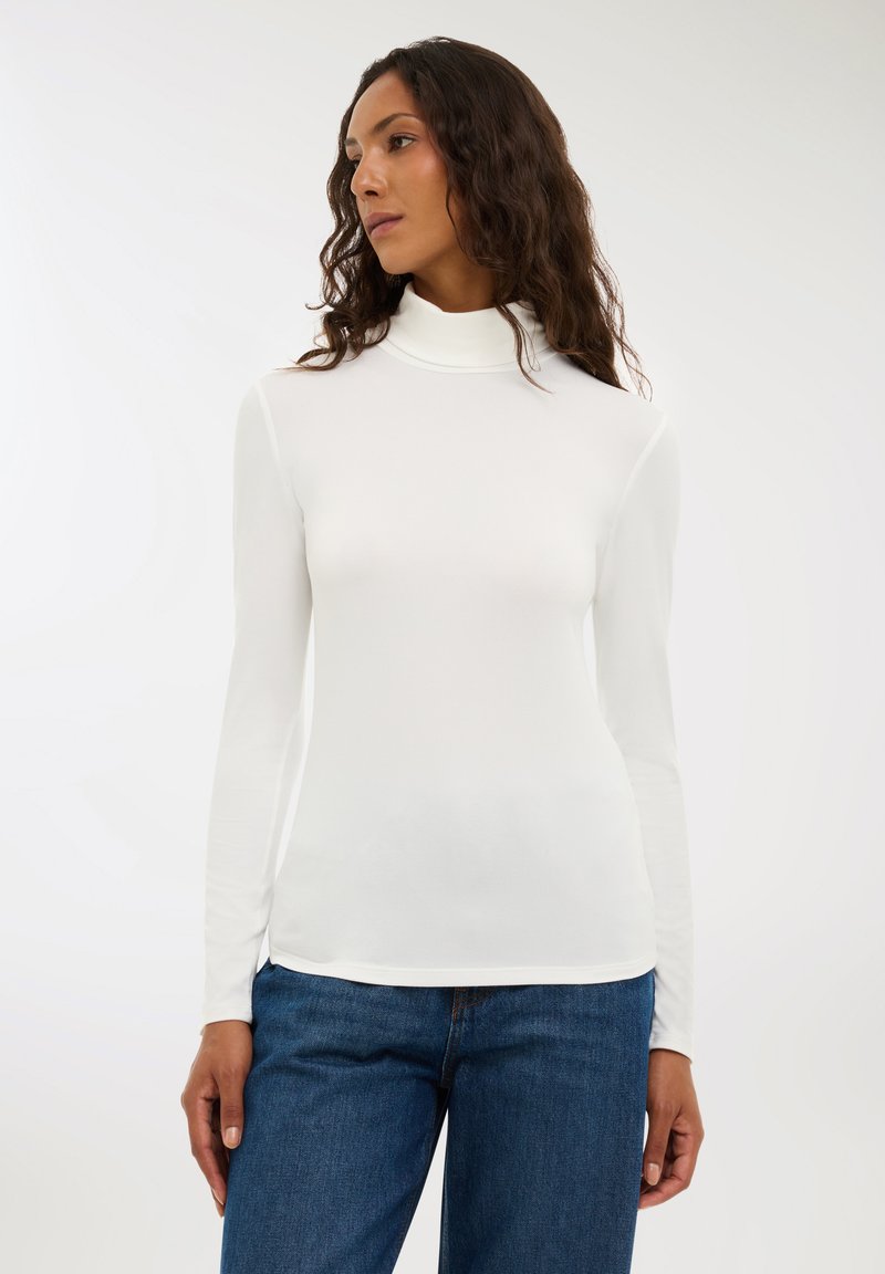 Maglia a maniche lunghe bianca con collo a dolcevita, realizzata in materiale morbido, caratterizzata da una silhouette aderente e una trama liscia. Indossata con jeans di denim blu.