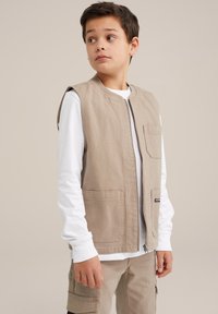 Beige ärmlös väst med dragkedja fram, två stora framfickor och en rund krage, bärs över en vit långärmad skjorta.