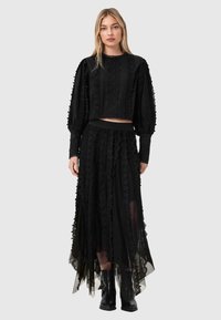 Γυναίκα με μαύρο δαντελένιο μπλουζάκι με μακριά μανίκια και αντίστοιχη φούστα με στρώσεις, φορώντας μαύρες μπότες, στέκεται μπροστά σε απλό φωτεινό φόντο.