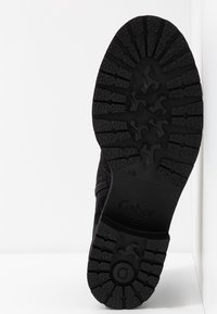 Semelle en caoutchouc noir avec motif de bande de roulement texturé, comportant des zones surélevées pour l'adhérence et un logo de marque. Les côtés de la chaussure sont visibles en tissu doux.