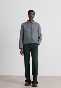 Casaco cinza em mistura de lã com colarinho e botões à frente, combinado com calças verdes. O conjunto apresenta um corte solto e detalhes de design sutis.